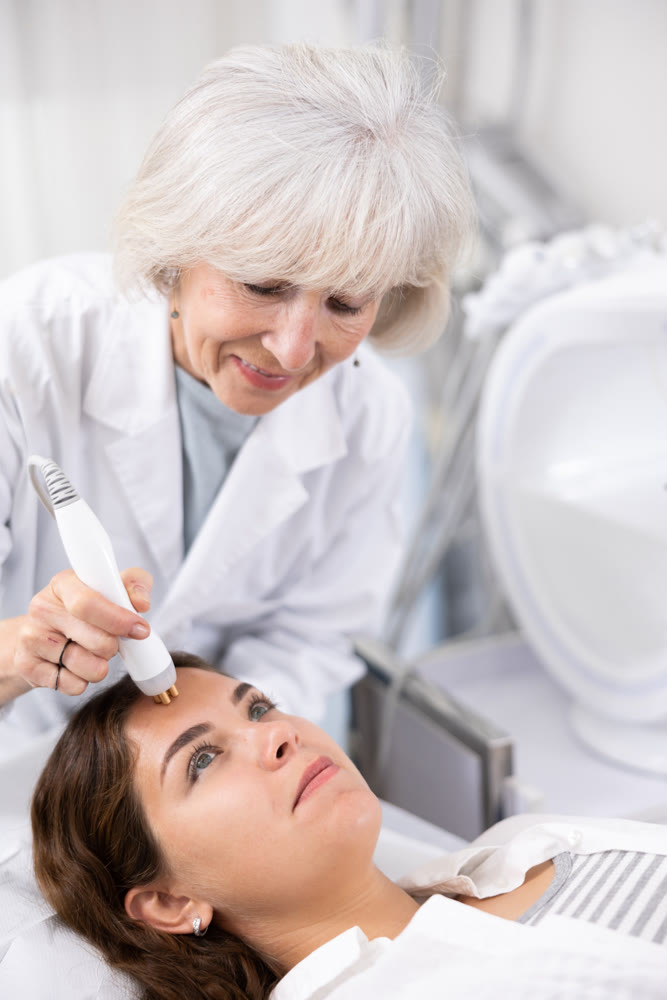 Mature female dermatologist performing cosmetology procedure for young woman, RF lifting and vacuum massage for facial skin therapy in clinic