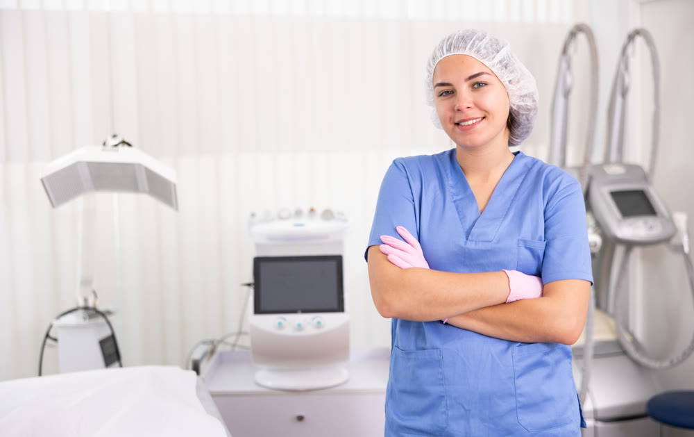 Young woman cosmetologist standing with crossed hands, smiling and looking at camera in clinic of aesthetic medicine