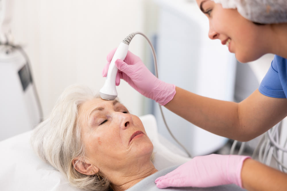 Professional young female cosmetologist doing facial skin ultrasound cavitation for for face lifting of mature woman laying in beauty salon