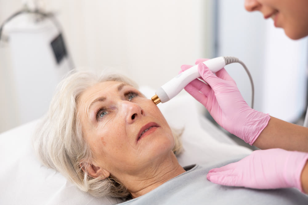 Young beautician doctor doing RF lifting procedure for face lifting of mature woman laying in beauty salon. Hardware cosmetology