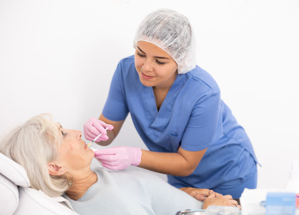 Professional young female beautician with syringe injecting filler with hyaluronic acid in lips of mature woman client during procedure in beauty salon