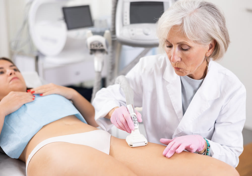 Elderly female doctor doing Rf lifting procedure on legs, buttocks and hips of young woman in beauty salon. Treatment of flabby skin