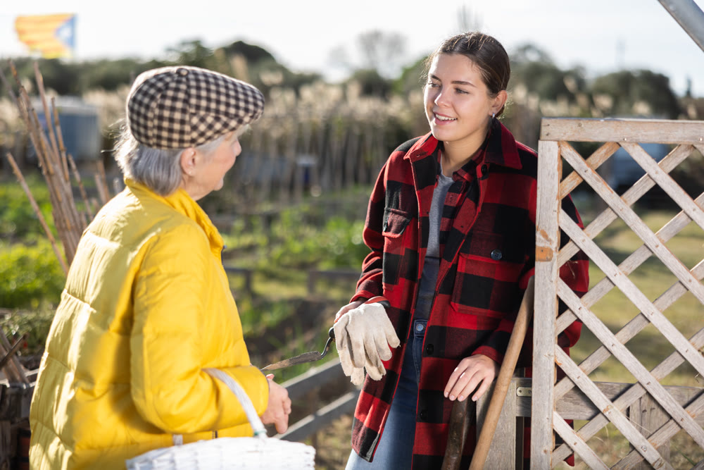 Two cheerful young and old female discussing gardening while standing near fence on garden during daytime in September