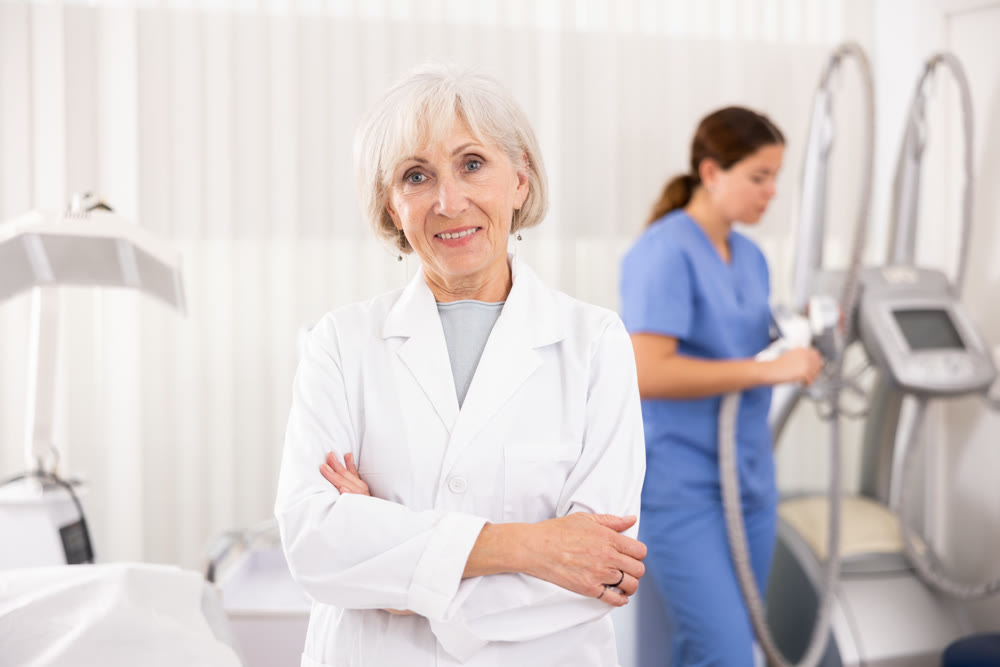 Smiling old woman doctor posing against background of cosmetology devices