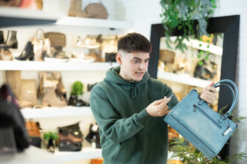 Shocked guy looking at price tag on womens handbag in trendy boutique