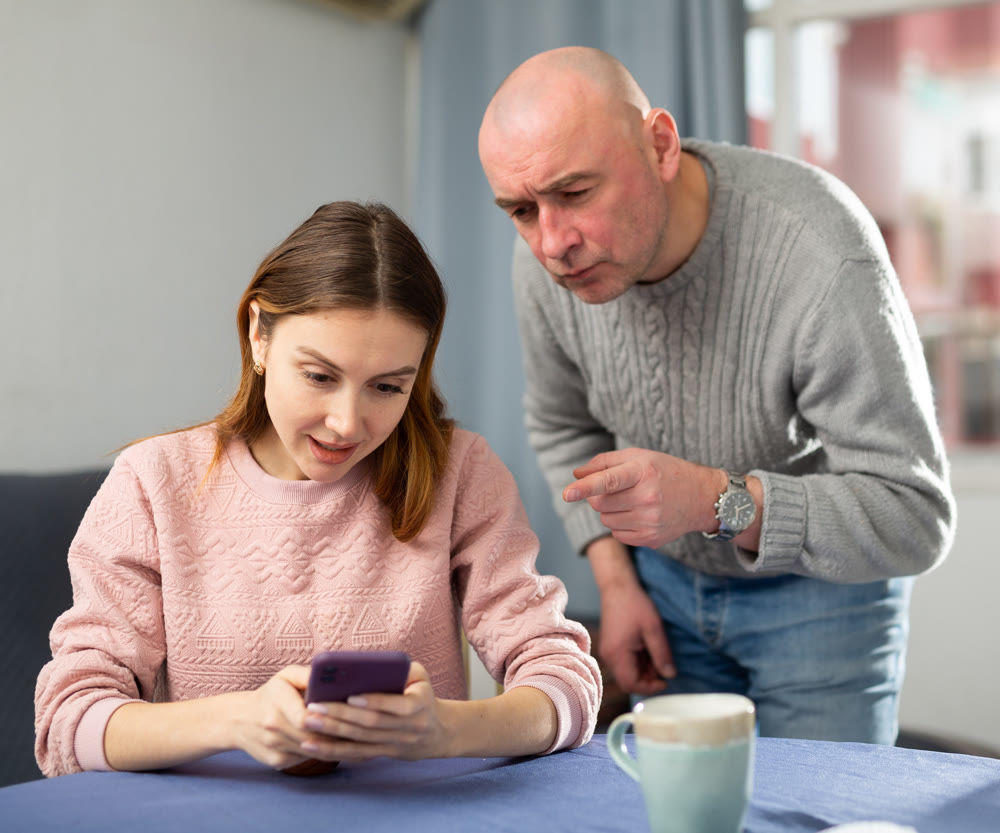 Angry husband observing his wife sitting and using phone