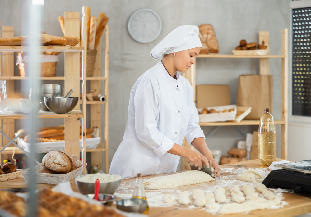 Female baker in uniform cutting raw dough with knife to make croissants or buns