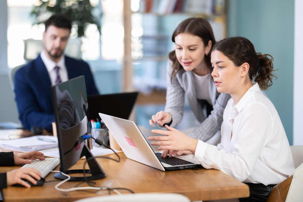 Young woman helping female colleague work on laptop
