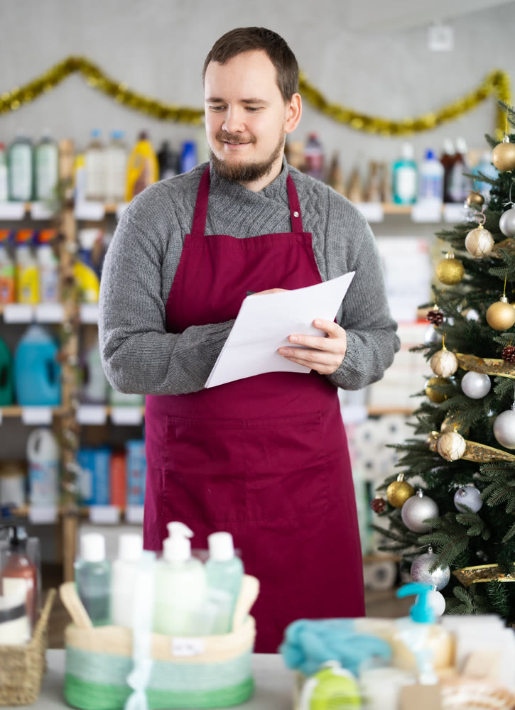 Attentive male salesperson restock of goods, checking papers in small household store decorated for New Year celebration
