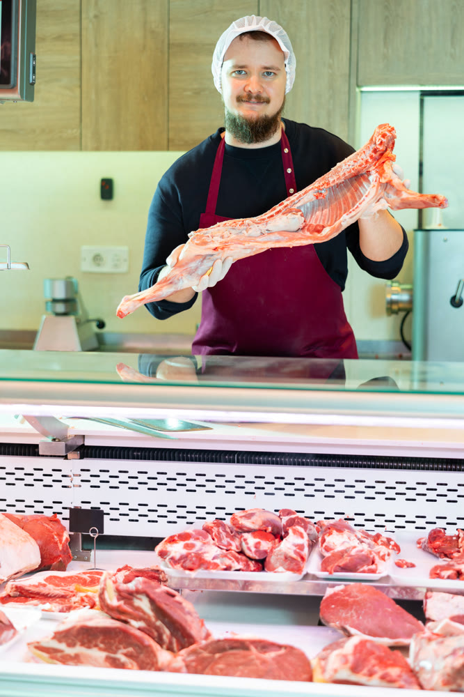 Bearded butcher demonstrating half of fresh lamb carcass at counter