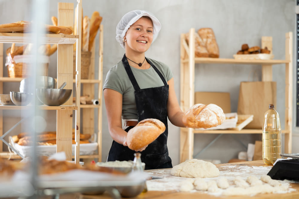 Young woman baker are standing near product range in bakery kitchen with basket of bread.