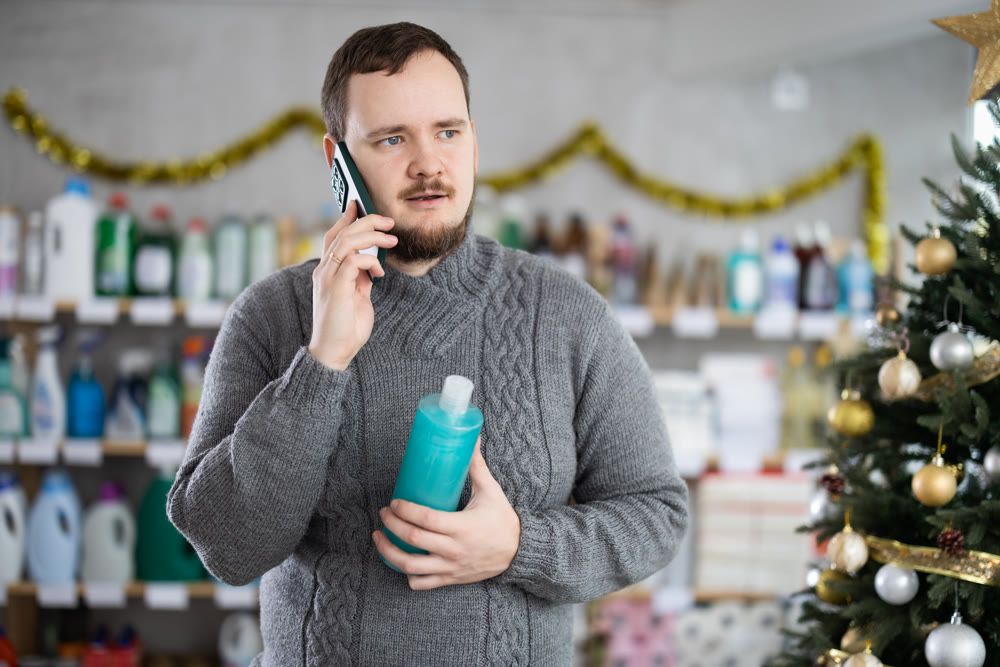 Handsome man speaking mobile and choose something in cosmetics store decorated for New Year holiday