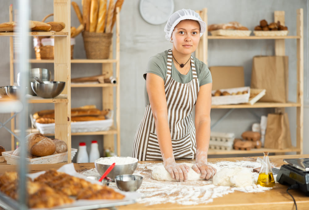 Young smiling female baker preparing raw yeast dough on kitchen table in bakery