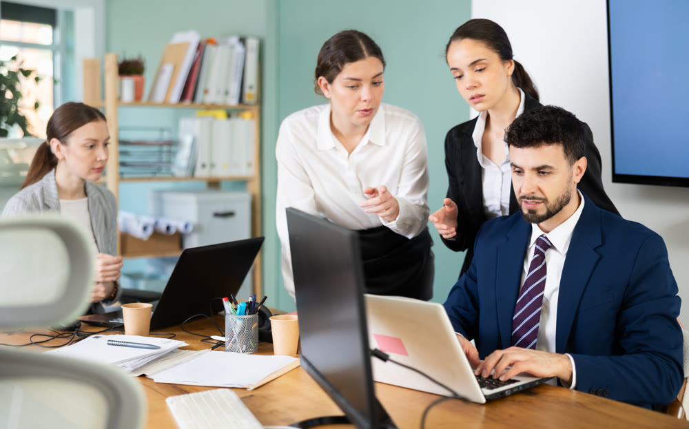 Two women help male colleague work on laptop
