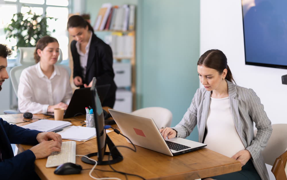 Pregnant woman is working on a laptop in an office while colleagues are discussing a project