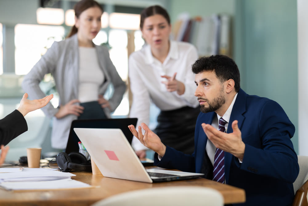 Frustrated man sits at a table with a laptop in front of his colleagues in office