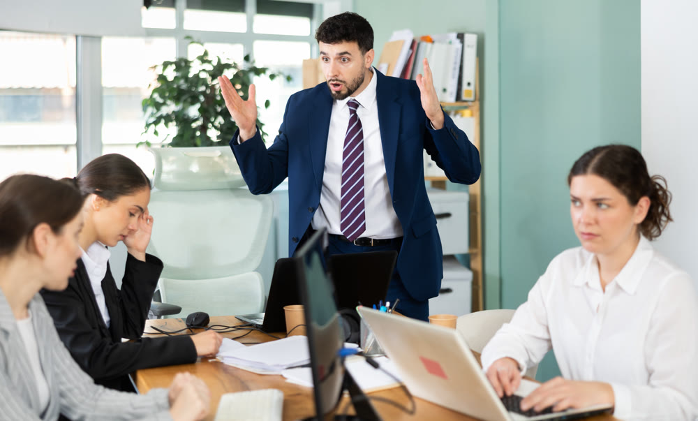 Young man standing and scolding sad seated colleagues in office