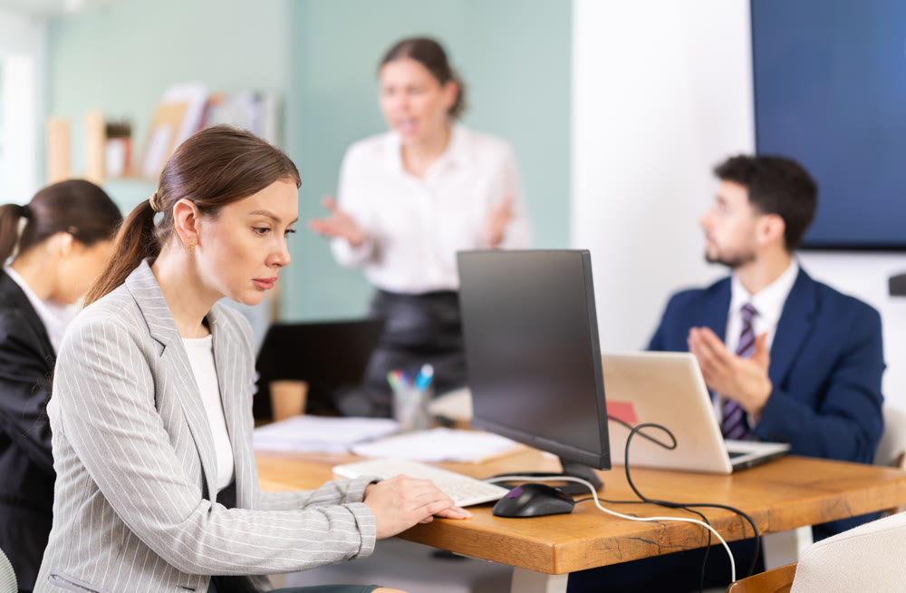 Upset female employee sitting at desk in front of team