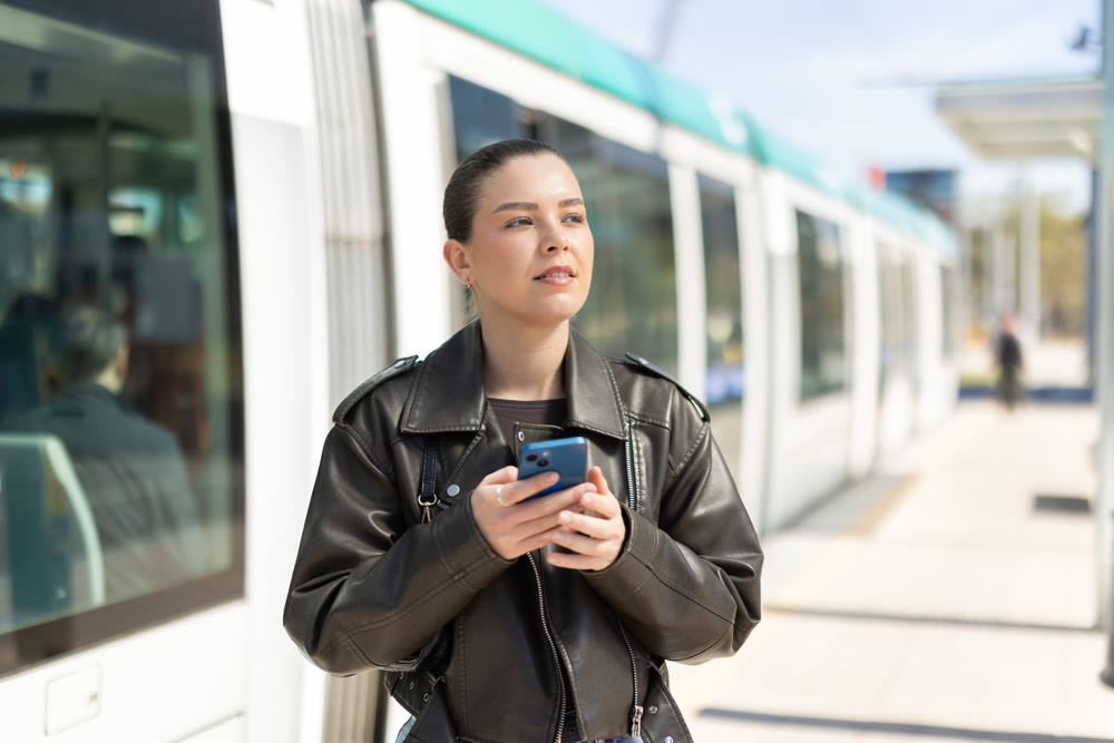 Portrait of girl in black jacket with mobile phone in her hands at tram stop on sunny spring day