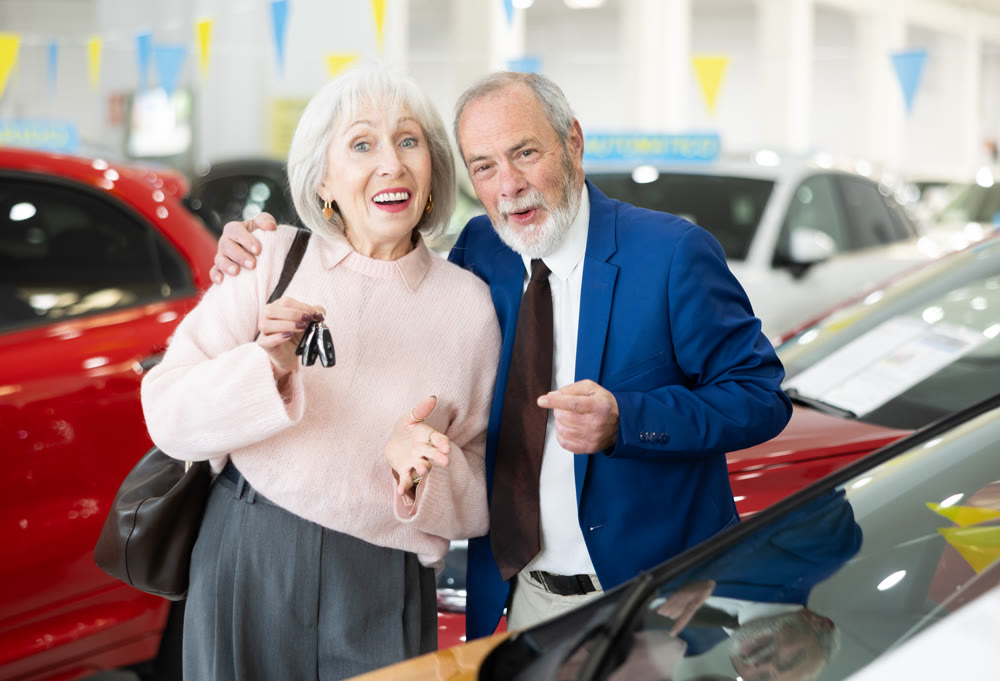 Joyful elderly couple stands near the car with a set of keys after purchase.