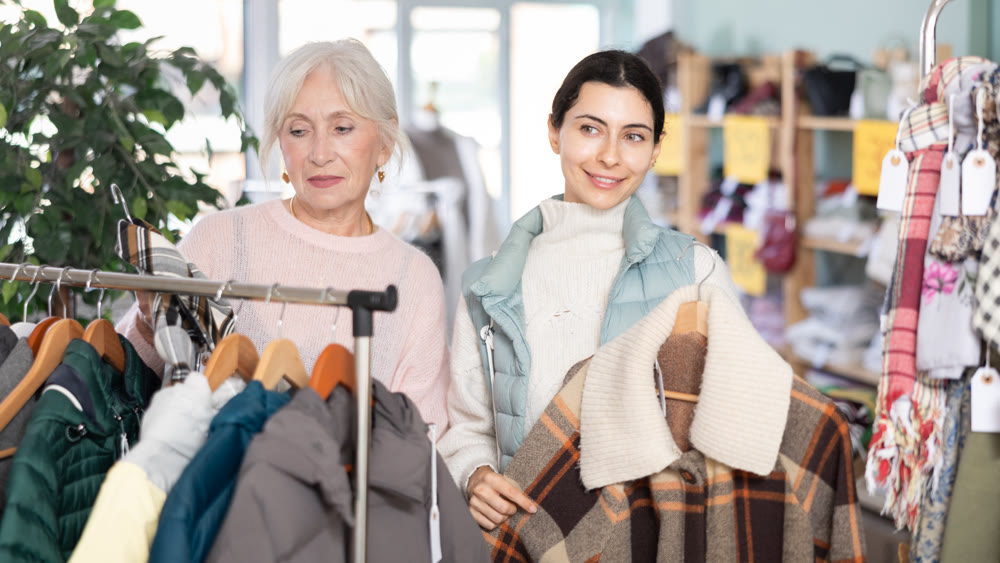 Two interested young and mature women standing in clothing shop and looking new coat or jacket