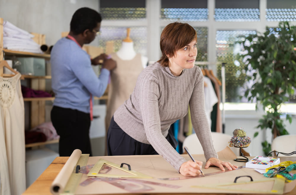 Woman draw dress pattern of product on paper. Companion work in background with mannequin