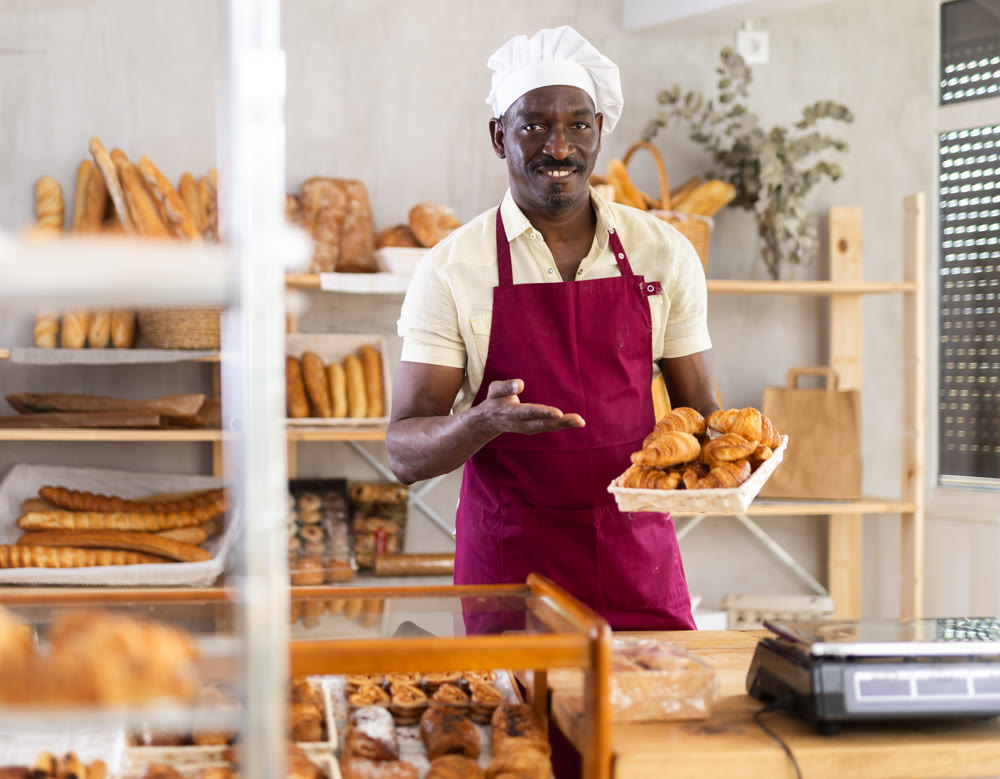 African male baker worker sells croissants, shows many products