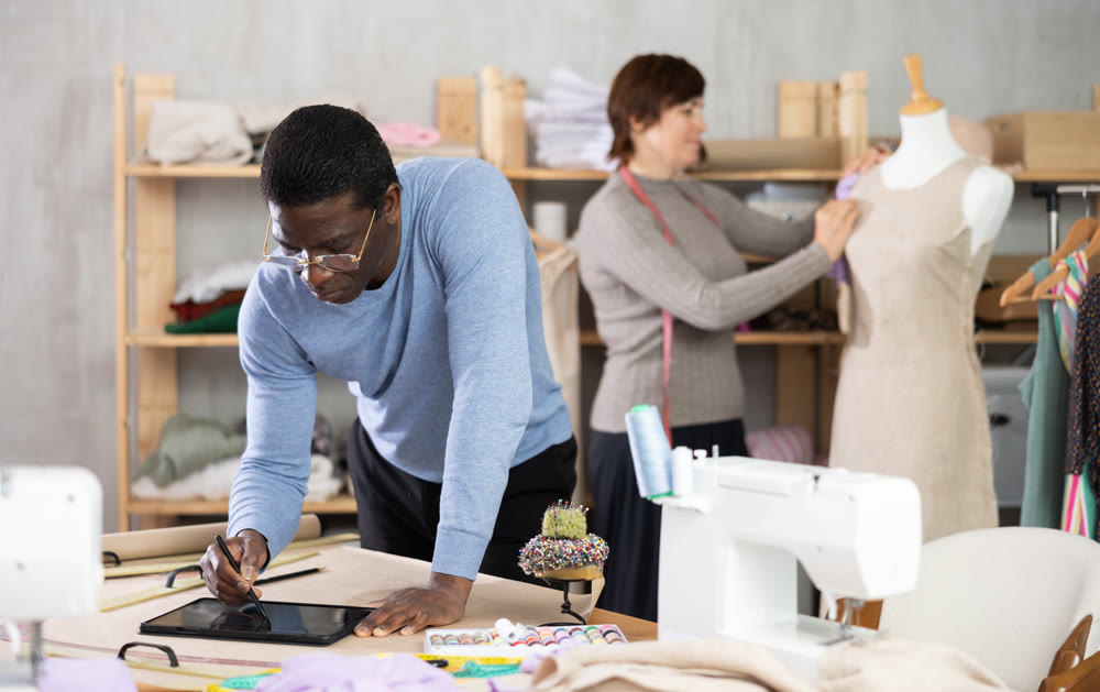 Fashion designer sketching garment on tablet while colleague working with mannequin