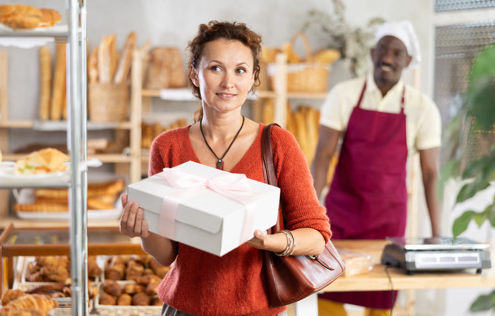 Woman buyer with pastry and baguettes in paper package stands near glass showcase in bakery