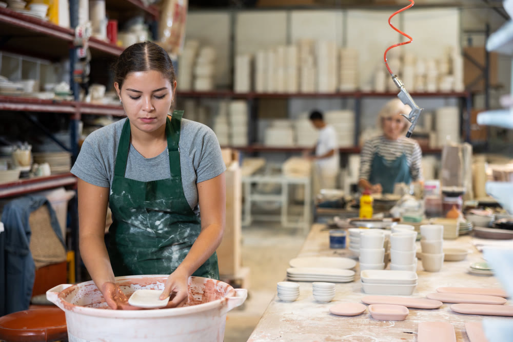Young female worker in pottery factory lowers fired clay plates and cups into container with glaze