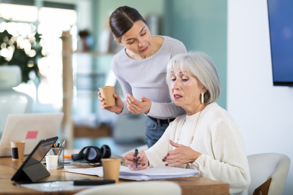 Elderly woman taking notes in document adult colleague helps
