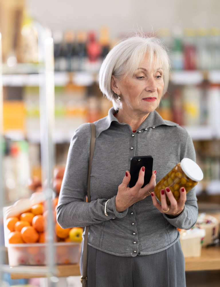 Checking expiration date of canned green olives - elderly female shopper scans QR code on label of jar using smartphone