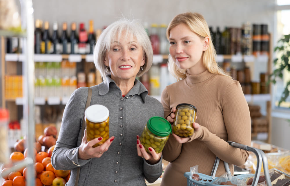 Girl and grandmother buyers examine good, doubt and choose pickled olives in store