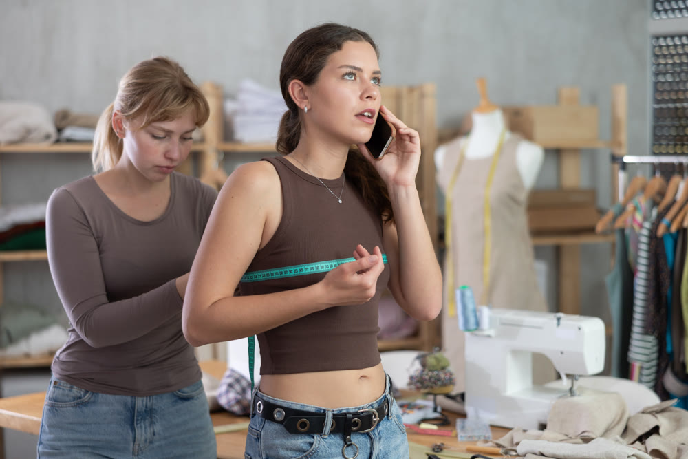 Young tailoress measuring female client speaking on mobile
