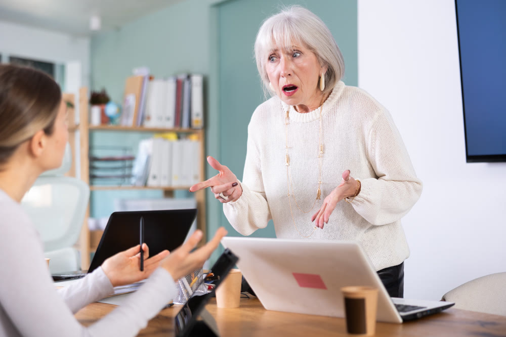 Female mature boss yells at woman employee, staff work at tablet