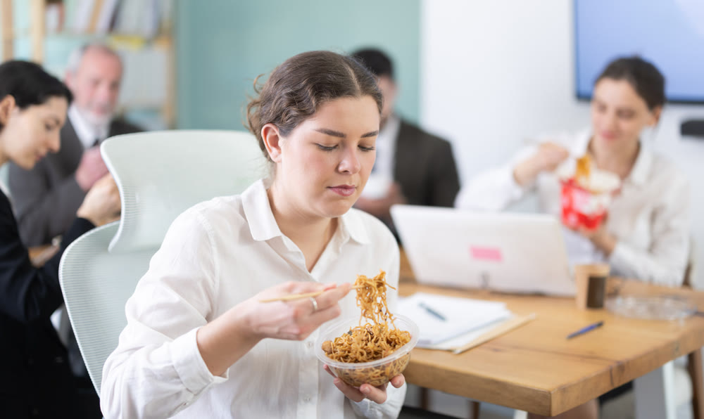 Young beautiful businesswoman having takeout meal during lunch break at work sitting at workplace at laptop with other colleagues in office