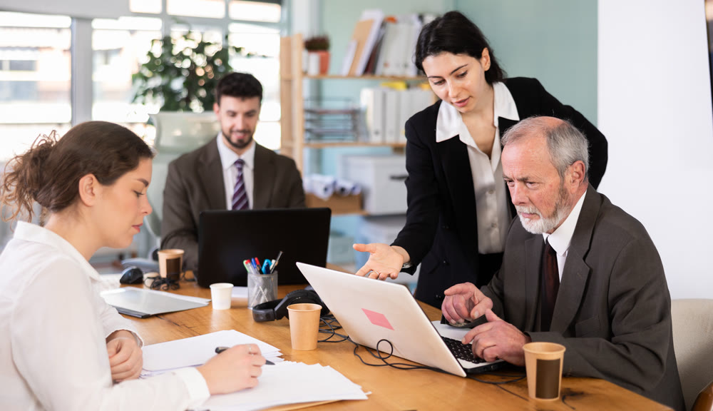 Girl leaned over to seated senior man employee, helps with laptop work