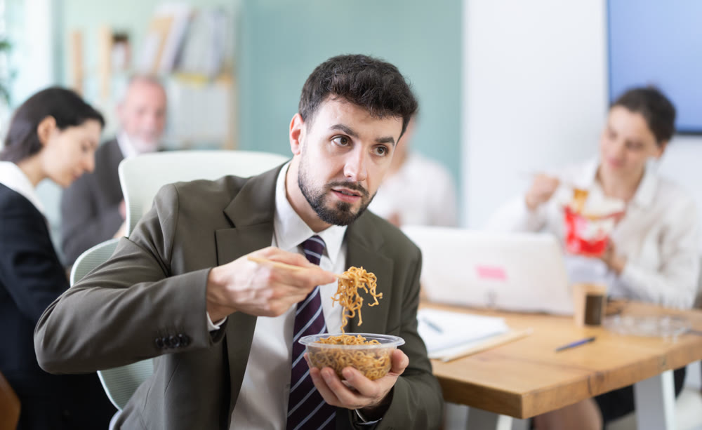 Young businessman having takeout meal during lunch break at work sitting at workplace at laptop with other colleagues in office
