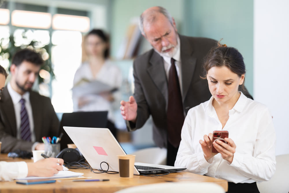 Strict mature man boss scolding girl office worker for scrolling social media on smartphone during work hours in office