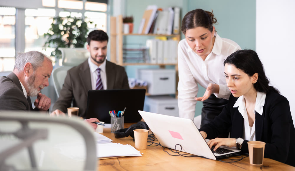 Focused young girl business worker helping businesswoman colleague with laptop, discussing new project in office