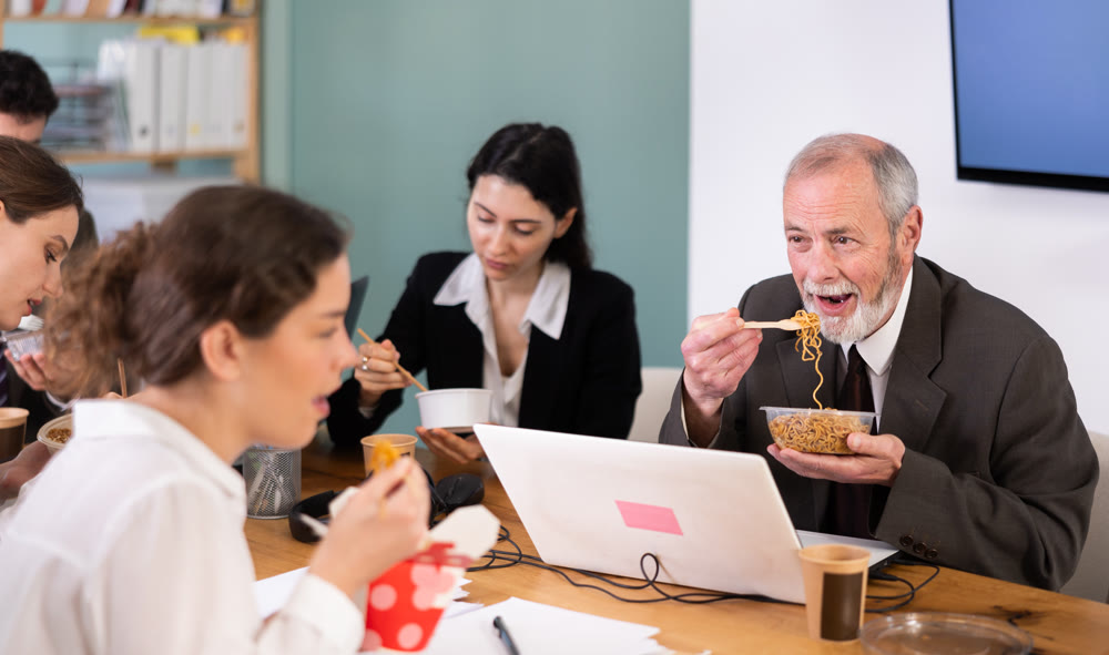 Office workers resting on break together eating, drinking coffee and talking, sitting at workplace at table with documents