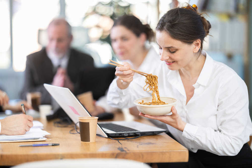 Positive young female office worker eating food at workplace, while other co-workers unhappy and continue to work in bright office of business company