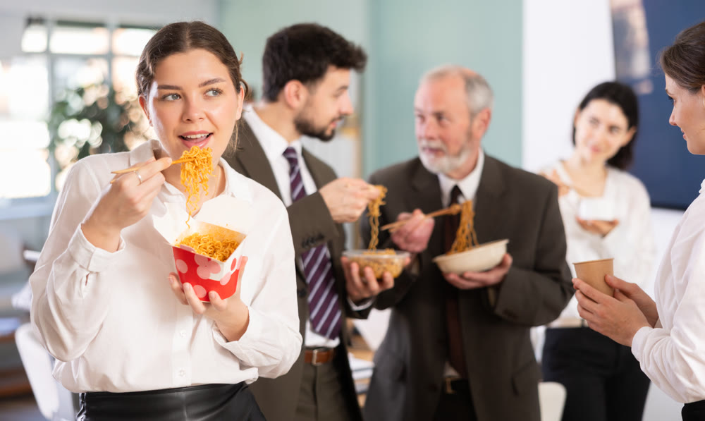 Happy girl office worker standing and eating fast food at office canteen with other colleagues