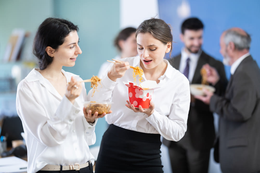Happy two businesswomen eating food together during lunch time in office of business company