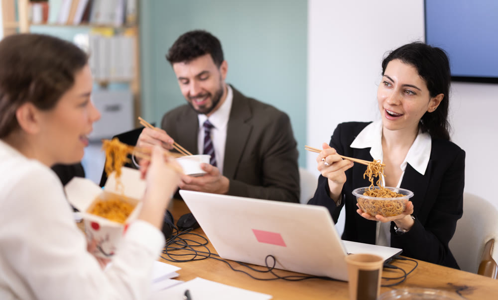 Office workers resting on break together eating, drinking coffee and talking, sitting at workplace at table with documents