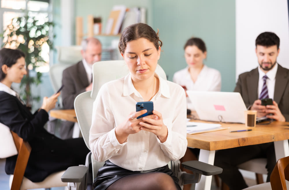 Positive female office worker scrolling smartphone sitting at workplace in office of business company