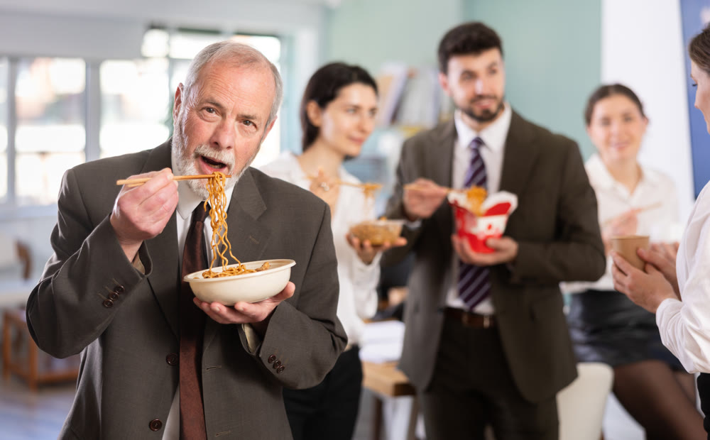 Mature businessman having takeout meal during lunch break at work with other colleagues in office