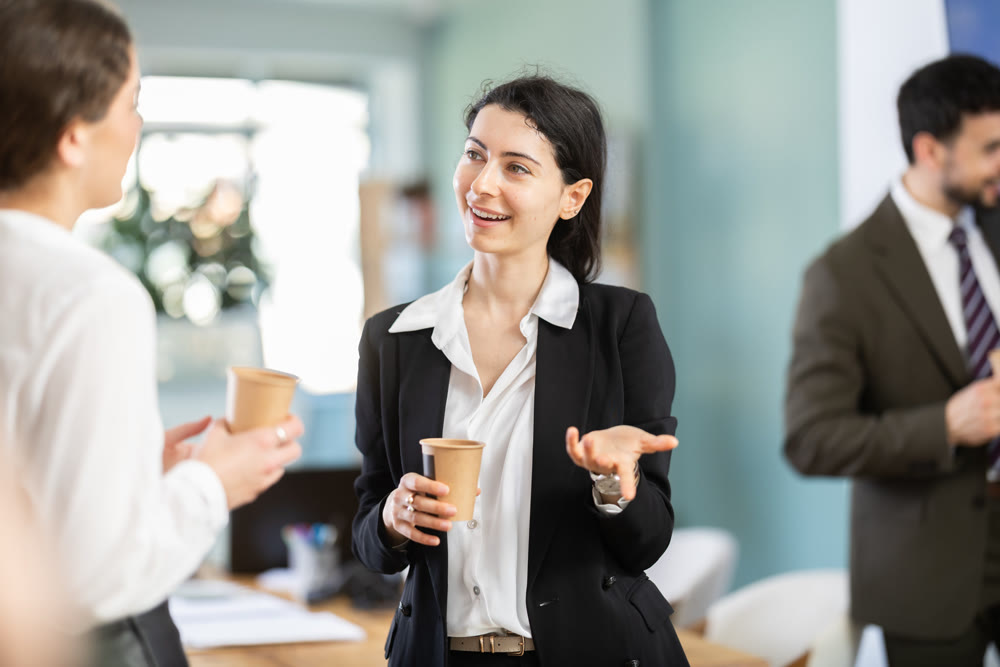 Two young woman co-workers employees drinking hot coffee and talking during break while working in office
