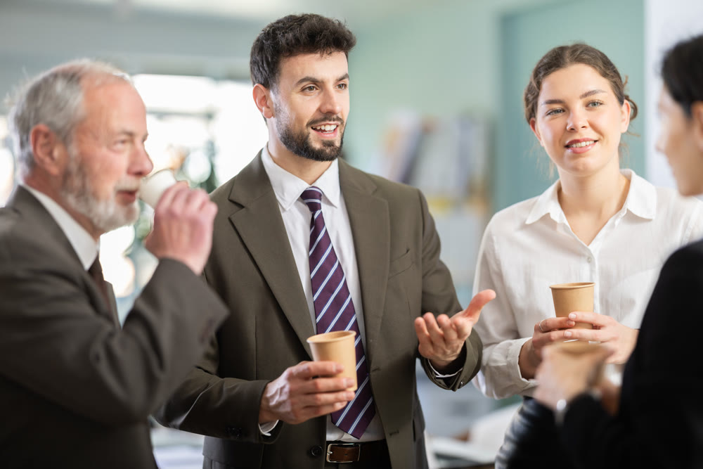 Group of positive office workers drink coffee and chat during work break in office of business company