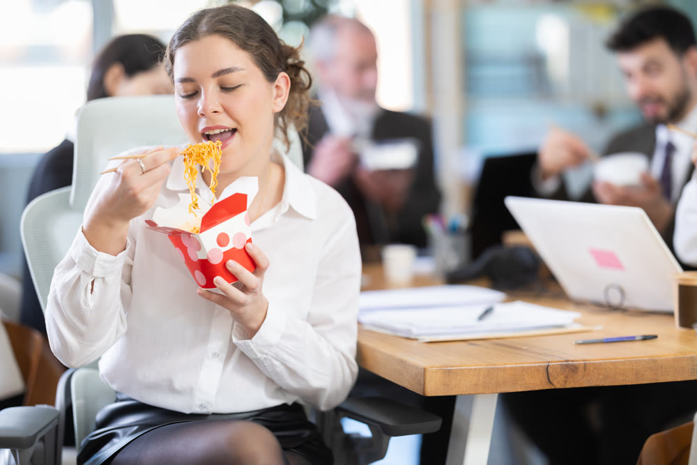 Young beautiful businesswoman having takeout meal during lunch break at work sitting at workplace at laptop with other colleagues in office